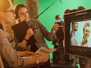 Female Director in chair looks at display and talks with Assistant, shooting historical film with green screen.