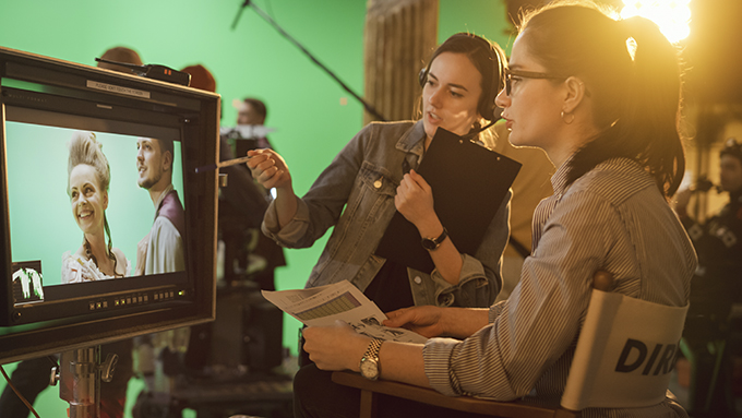 Female Director in chair looks at display and talks with Assistant, shooting historical film with green screen.