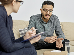 Young mixed-race man wearing eyeglasses sitting on cozy sofa and talking with private practice substance abuse counselor.