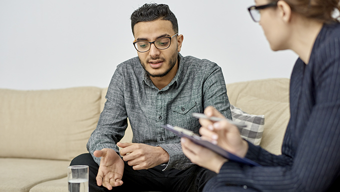 Young mixed-race man wearing eyeglasses sitting on cozy sofa and talking with private practice substance abuse counselor.